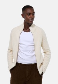 Cardigan en tricot beige avec un col montant, fermeture �éclair intégrale, texture côtelée et manches longues, associé à un t-shirt blanc et un pantalon marron.