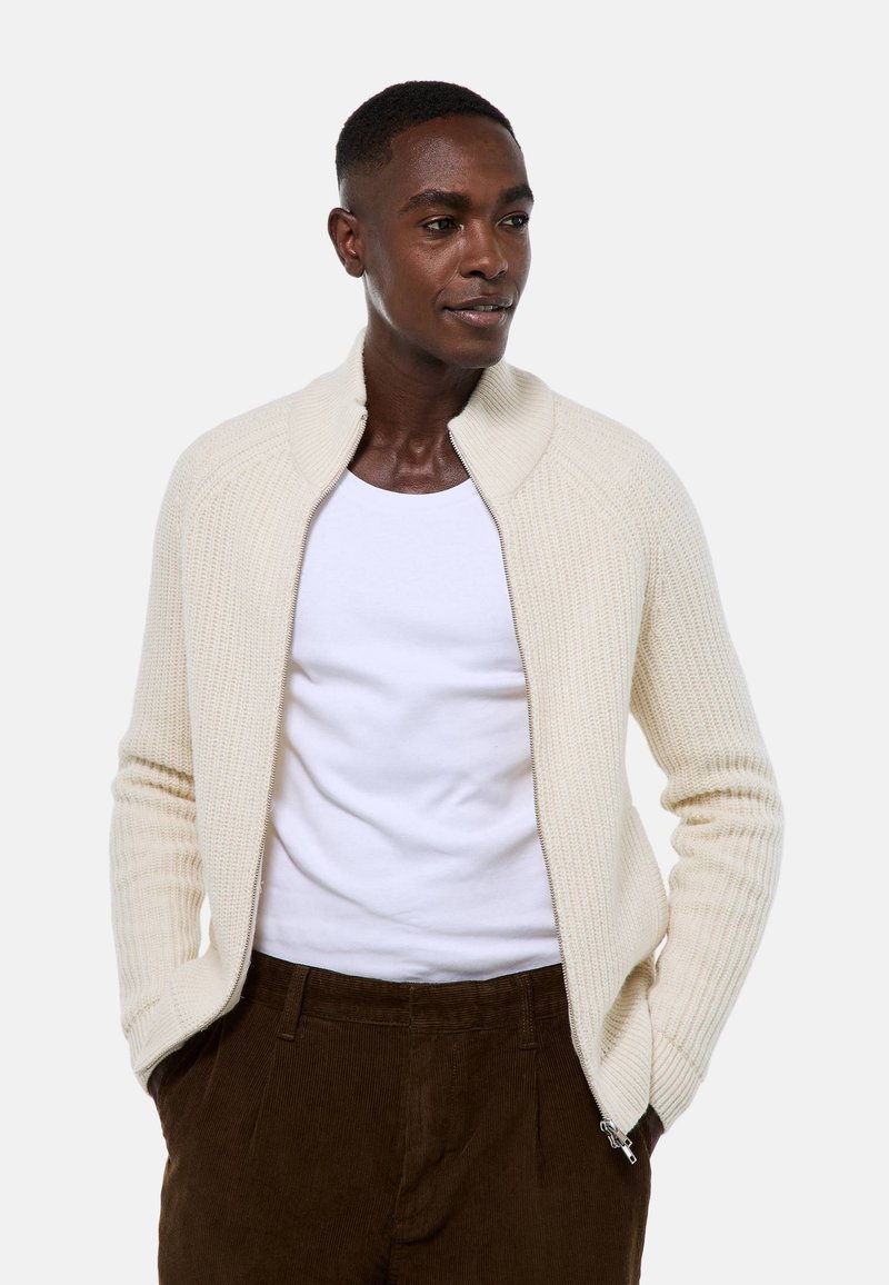 Cardigan en tricot beige avec un col montant, fermeture éclair intégrale, texture côtelée et manches longues, associé à un t-shirt blanc et un pantalon marron.