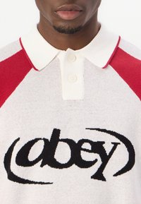 Polotröja i vitt med röda raglanärmar, med en framträdande svart "obey"-logotyp på framsidan, tillverkad av stickad tyg.