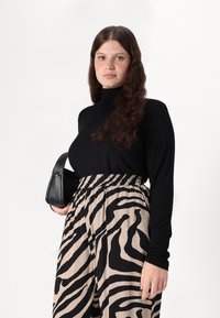 Must mustreväga mustade pikkade varrukatega pluus, millele on sobitatud beežid püksid, millel on must zebra triipude muster. Modell hoiab käes väikest musta käekotti.