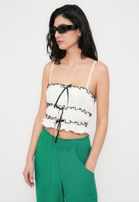 Haut blanc court avec des volants et des liens noirs, associé à un pantalon vert à jambes larges avec poches. Porté avec des lunettes de soleil noires.