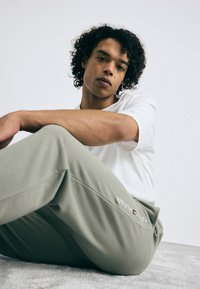 Lys grå afslappede sweatpants med et broderet logo, parret med en simpel hvid t-shirt. Model siddende på en tekstureret overflade.