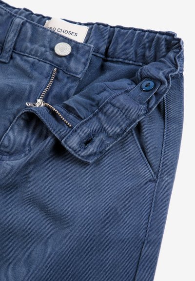 Pantalon bleu en coton avec taille élastique, fermeture éclair partiellement ouverte, rabat boutonné et une étiquette blanche pliée à l'intérieur de la ceinture.