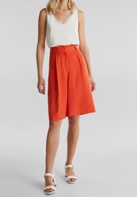 Bílý bezrukávový top s výstřihem do V, kombinovaný s oranžovými culottes s vysokým pasem. Modelka má na sobě bílé sandály na pásku. Hladká textilie.