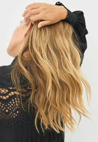 Długie, faliste blond włosy opadają na czarną bluzkę z misternymi koronkowymi detalami na ramionach i mankietach.