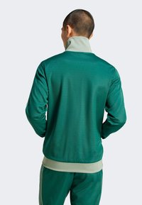 Grön sportjacka med hög krage, tillverkad av slät tyg; har kontrasterande ljusgröna detaljer vid kragen och fållen.