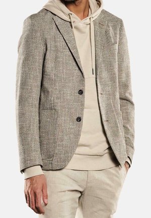 Mężczyzna ubrany w beżową, teksturowaną marynarkę na jasnobeżowej bluzie z kapturem i beżowych spodniach, stojący z jedną ręką w kieszeni na białym tle.