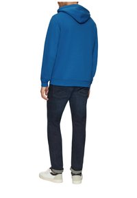 Blauwe hoodie met een buidelzak, gecombineerd met donkerdenim jeans en witte sneakers. De stof lijkt glad en dik te zijn.