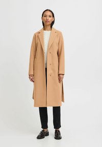 Manteau en mélange de laine couleur camel avec une coupe ajustée, un devant simple boutonné, des revers en notch et des poches latérales. Associé à un pantalon et des chaussures noirs.
