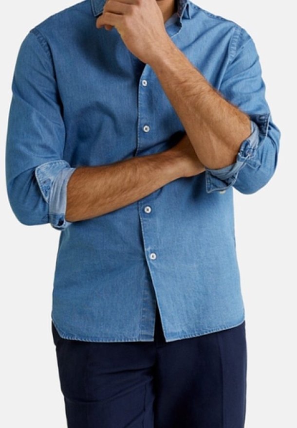 Camisa de mezclilla azul claro con cierre de botones al frente, mangas largas remangadas y textura sutil. Combinada con pantalones de color azul marino.