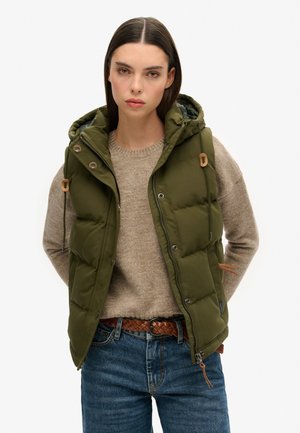 Olivgrüner Steppwesten mit Kapuze, Druckknöpfen und Kordelzügen, über einem beigen Pullover getragen. Das Modell trägt dunkle Jeans.
