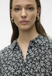 Chemise noire à motifs floraux blancs, dotée d'un col, de boutons et d'une texture de tissu léger. Gros plan sur le visage du mannequin.
