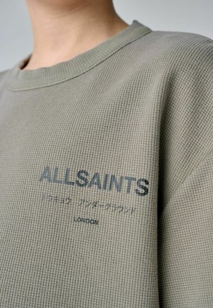 Detailní záběr osoby oblečené v olivově zelené texturované košili s nápisem "ALLSAINTS" a japonskými znaky na hrudi.