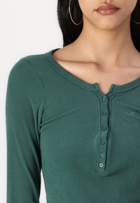Grønn, stripete langermet topp med rund hals, knappestolpe med seks knapper, og subtil kontrastlogo-detalj.