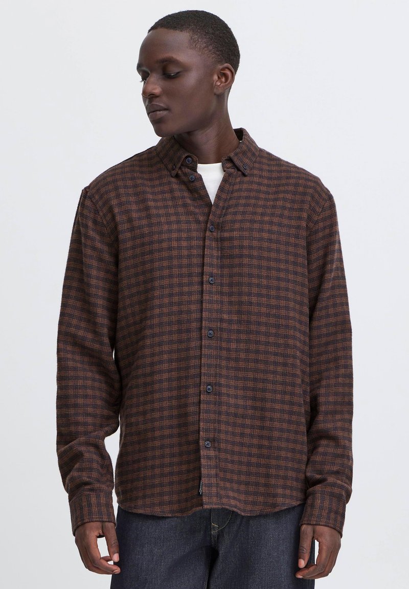 Homme portant une chemise à carreaux marron et noire par-dessus un t-shirt blanc et un jean foncé, regardant sur le côté devant un fond uni.