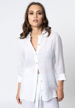 Vrouw met golvend bruin haar, die een wit, knoopgesloten overhemd met lange mouwen en bijpassende witte broek draagt tegen een effen achtergrond.