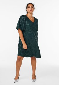 Abito verde con paillettes, scollatura a V e maniche a sbuffo corte, caratterizzato da una vestibilità comoda e una texture scintillante, abbinato a scarpe argento con punta.