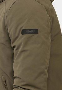 Giacca verde oliva con tessuto liscio, con una toppa logo nera sulla manica sinistra e dettaglio con cerniera sul davanti.