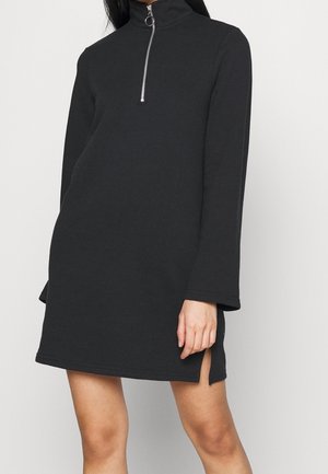 Frau in einem schwarzen Mini-Kleid mit langen Ärmeln, hohem Kragen und vorderem silbernem Reißverschluss, vor einem schlichten hellen Hintergrund stehend.