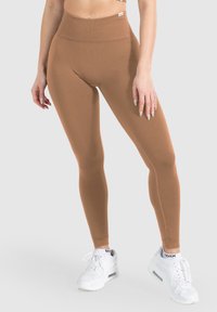 Bruine high-waisted legging gemaakt van rekbare stof. Heeft een gladde textuur en een strakke pasvorm, gecombineerd met witte sportschoenen.
