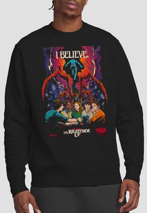 Sweatshirt mit fünf Teenagern, die ein Brettspiel spielen, mit einem großen roten Spinnenmonster und einer schwebenden Figur darüber, mit den Texten "I BELIEVE" und "The Right Side Up".