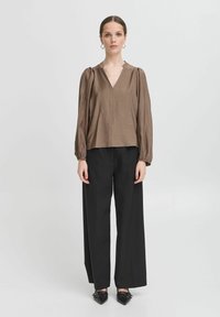 Blusa marrone, strutturata, con scollatura a V e maniche a sbuffo, abbinata a pantaloni neri a gamba larga e scarpe con punta. Design semplice e moderno.