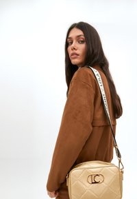 Una giovane donna con lunghi capelli scuri guarda oltre la spalla, indossando una giacca di camoscio marrone e portando una borsa in oro con un logo.