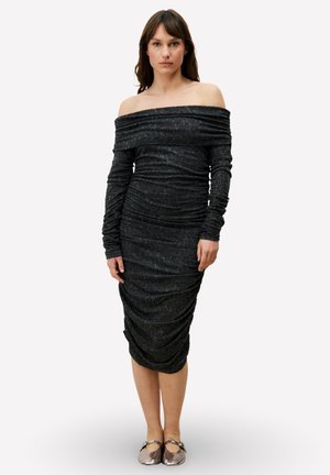Robe noire épaules dénudées avec détails froncés, manches longues et tissu texturé, longueur jusqu'aux genoux, associée à des chaussures plates métalliques.