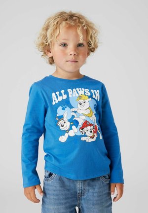Blond child wearing blue long-sleeve shirt with "ALL PAWS IN" text and three cartoon puppies in police, firefighter, and construction hats.