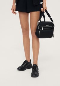 Černá kabelka se dvěma zipovými přihrádkami, vyrobená z měkké látky. Má odnímatelný popruh a kovové kování. Doplněna černými teniskami.