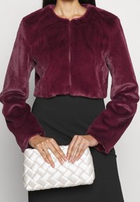 Veste en fausse fourrure bordeaux courte à manches longues, associée à une pochette matelassée blanche. Portée par-dessus une robe noire.