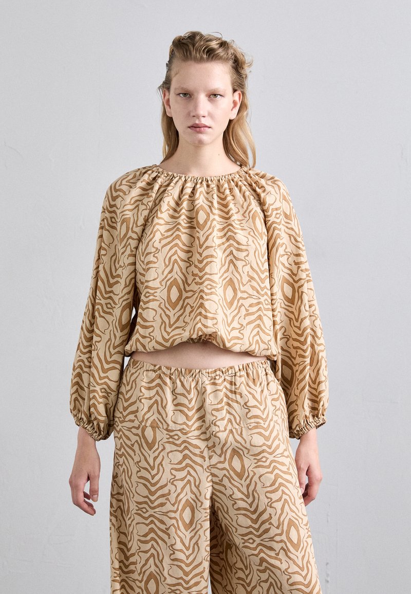 Beigefarbenes strukturiertes Outfit mit einem Cropped Top und weit geschnittenen Hosen mit einem wellenförmigen braunen Muster. Das Design umfasst geraffte Ärmel und einen gekräuselten Bund.