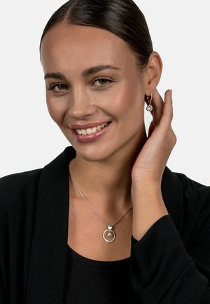 Silberne Halskette mit rundem Anhänger, der eine Perle zeigt. Passende Ohrringe mit runden Akzenten. Das Model trägt ein schwarzes Outfit.