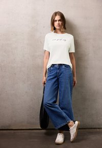 T-shirt crème à manches courtes avec le texte "new york city", associé à un jean bleu évasé et des baskets blanches. Sac noir porté sur l'épaule.