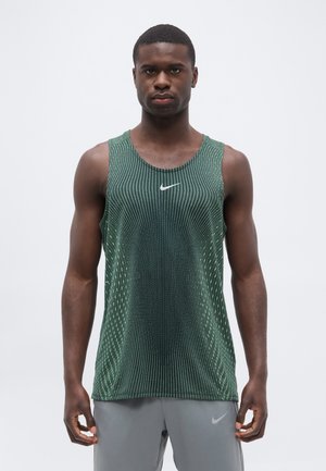 Homme portant un débardeur sportif vert sans manches de Nike et un short gris, debout devant un fond gris uni, face à l’avant.