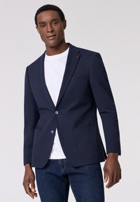 Blazer blu scuro con texture, dotato di due bottoni blu, revers a punta e tasche applicate, indossato su una maglietta bianca e abbinato a jeans scuri.
