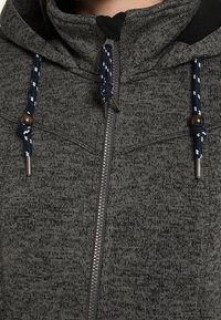 Šedá mikina na zip s texturovaným pletením. Má černý límec, modré a bílé šňůrky s dřevěnými doplňky a přední zip.