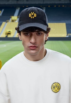 Junger Mann mit schwarzer BVB 09 Mütze und weißem Sweatshirt mit BVB 09 Logo, stehend im gelb-schwarzen Sitzbereich eines Stadions.