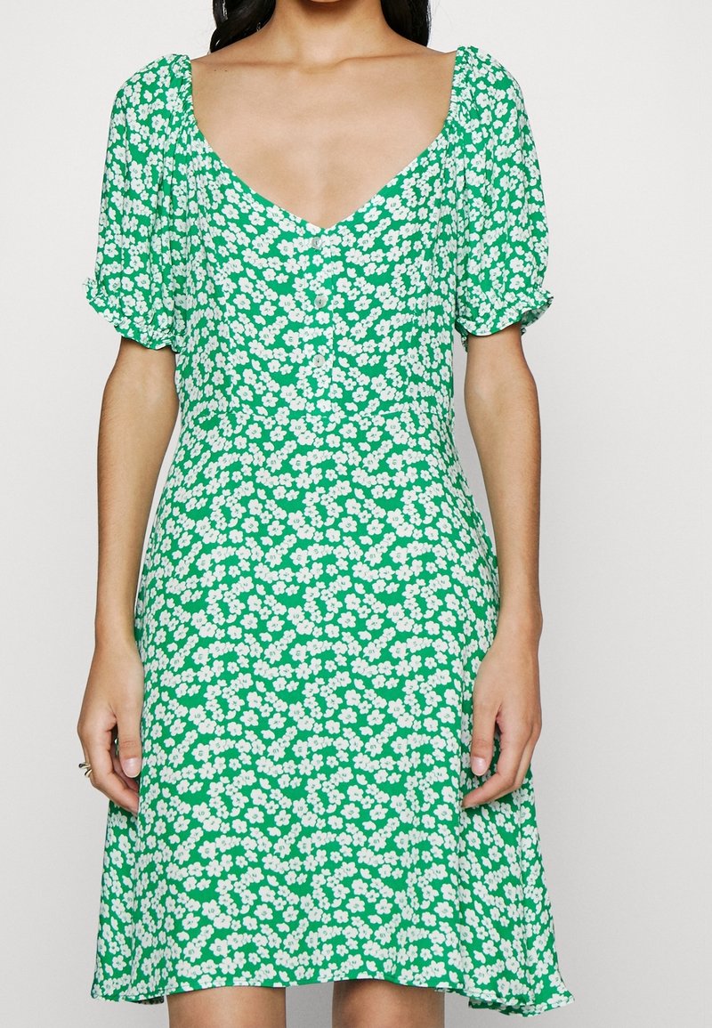 Femme portant une robe verte avec un motif floral blanc, des manches courtes bouffantes et un décolleté en V.