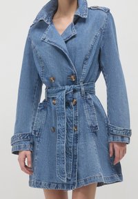 Denim trench coat in light blue, featuring a belted waist, double-breasted design with buttons, and large lapels. Textured fabric.