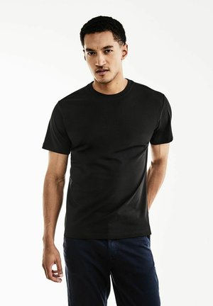 T-shirt noir à manches courtes en coton avec un col rond et une texture lisse. Porté avec un pantalon bleu marine, fond uni.