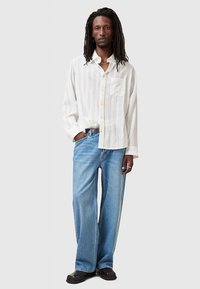 Weißes, gestreiftes Hemd mit Knöpfen und langen Ärmeln, kombiniert mit lockeren hellblauen Jeans und schwarzen Schuhen. Mit Taschen-Detail am Hemd.