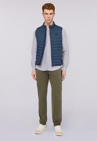 Chaleco acolchado azul con cuello alto, llevado sobre una camisa de botones gris claro. Pantalones verde oliva y zapatillas blancas completan el conjunto.