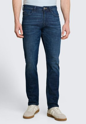 Mann trägt dunkelblaue Slim-Fit-Jeans und weiße Sneaker und steht vor einfachem Hintergrund.