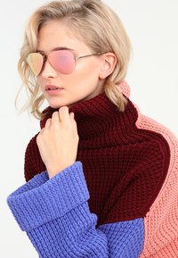 Pulôver com uma mistura de malha texturizada em bordeaux, rosa e azul. Modelo usa óculos de sol rosa reflexivos com armações de metal finas.