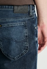 Sötétkék, enyhén kopott farmernadrág, texturált fekete bőrpírral a derékpánton és egy hátsó zseb díszvarrással.