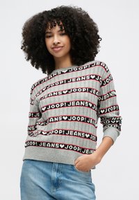 Mlada ženska s kodrastimi lasmi, oblečena v sivi pulover z napisom »JOOP! JEANS« in modre kavbojke, z roko v žepu, rahlo se smehlja.