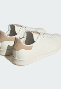 Hvide kanvas sneakers med en struktureret overflade, sandfarvede læderdetaljer ved hælen og en lys gummisål. Har seks snørrehuller og præget branding.