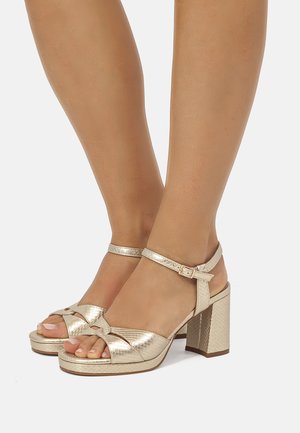 Pies con sandalias de tacón alto color dorado metálico, con tiras en el tobillo y un patrón texturizado tejido, sobre un fondo claro.