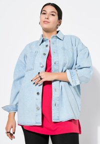 Veste en denim oversize bleu clair avec fermeture à boutons, poches sur la poitrine et manches retroussées, portée par-dessus une chemise rouge et un leggings noir.